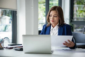 Businesswoman sitting at desk on couch in workplace or at home working on laptop and analyzing data on charts and graphs and writing on papers to make business plan and strategies for company