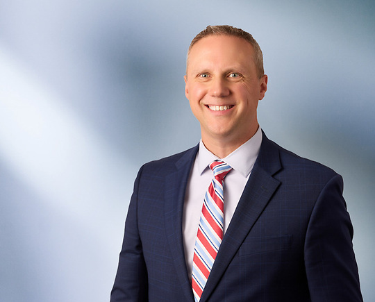 man, Todd Hoppe, with short light brown hair in a dark blazer, with a blurred blue and white background.